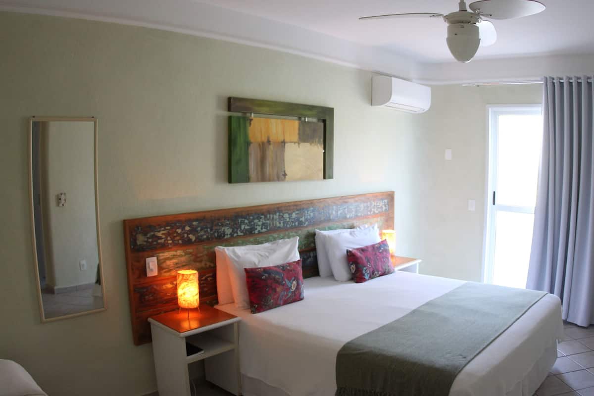 Una habitación de hotel con cama, mesitas de noche, televisor, ventilador de techo, ventana con cortinas y un cuadro en la pared.