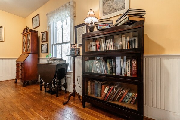 A room with a wooden floor, a desk with a lamp, a bookshelf filled with books, and a grandfather clock. - The Babcock House (Appomattox)