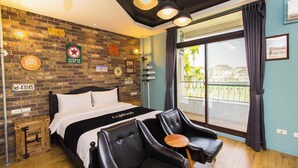 Quarto de hotel com parede de tijolos, cama com roupa de cama branca, poltronas de couro preto e uma janela grande com vista para prédios e palmeiras.