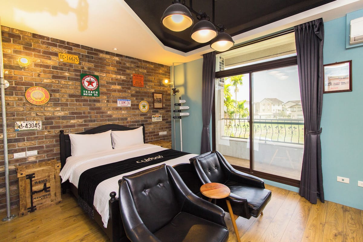 A hotel room with a brick wall, a bed with white bedding, black leather chairs, and a large window with a view of buildings and palm trees.