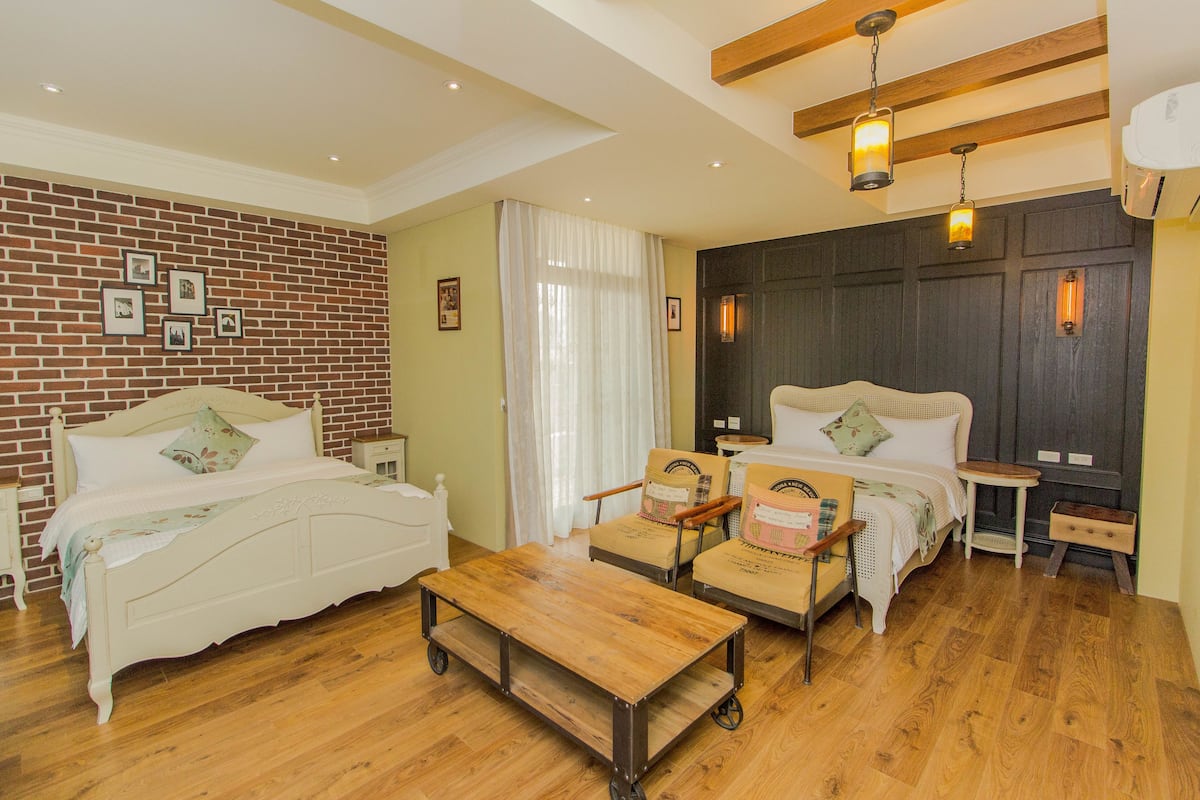 A room with a brick wall, two beds, a wooden coffee table, and a chair.