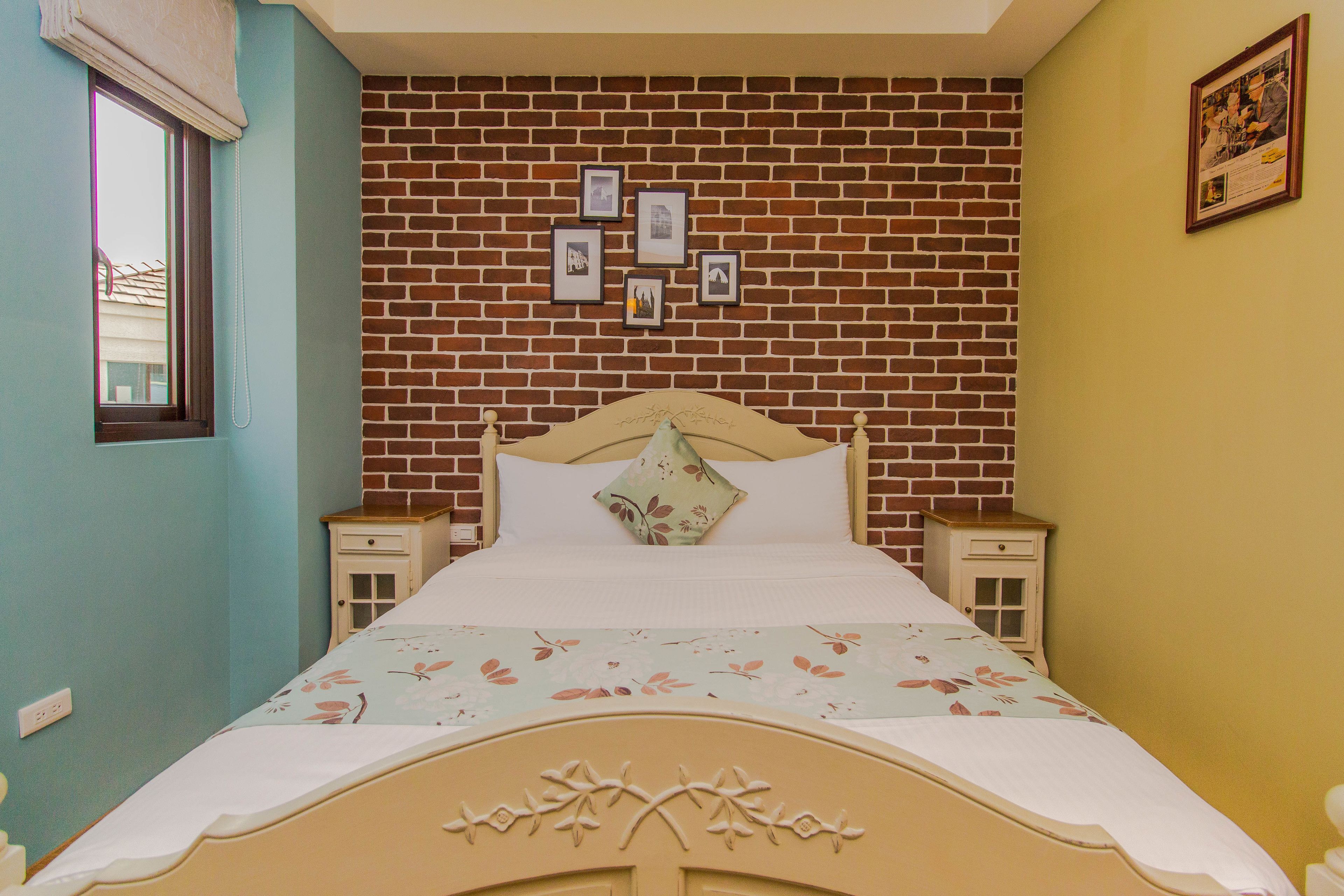 Une chambre avec un mur décoré d’un motif en briques, un lit recouvert d’une literie à motifs floraux et des tableaux encadrés accrochés au mur.