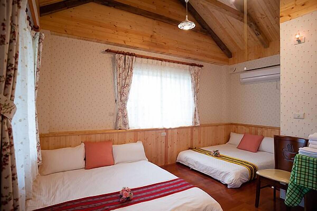 A room with two beds, wooden walls, a window with curtains, and a table with a chair.