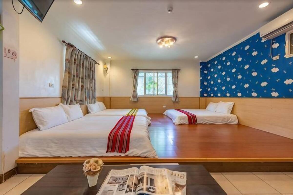 A hotel room with two beds, a window, a wall with a floral pattern, and a small table with a vase.
