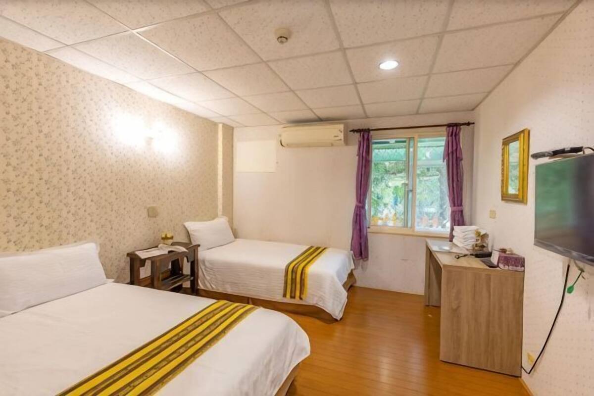 A hotel room with two beds, a desk, a chair, a window with curtains, and a television.