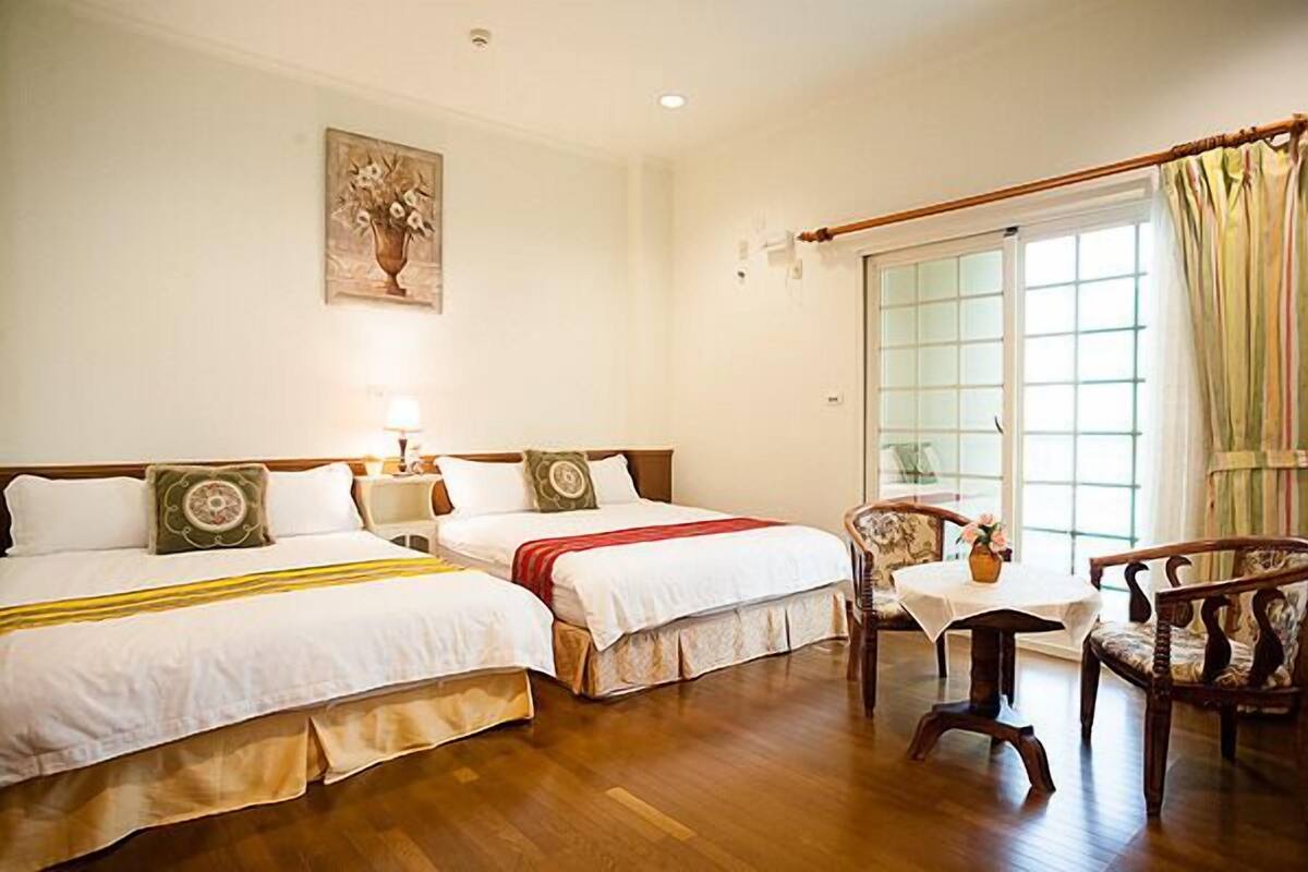 A hotel room with two beds, a wooden floor, a table with a vase, and a window with curtains.