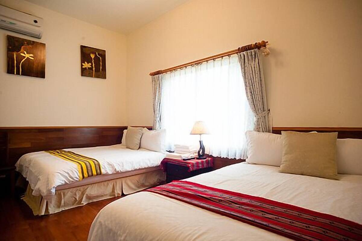 A hotel room with two single beds, a wooden headboard, a window with curtains, and two wall-mounted artworks.