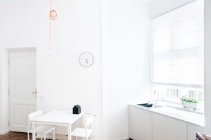 A bright, white room with a table and chairs, a clock, a window with blinds, and a radiator. - Boom Apartments (Kraków)