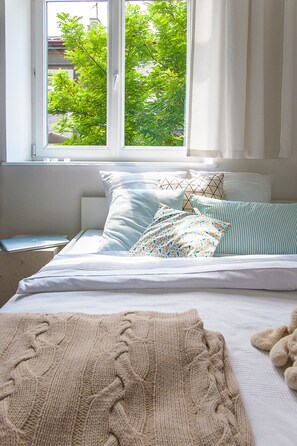 Una cama bien hecha con vistas a árboles por la ventana, una mesita de noche con una computadora portátil y una manta tejida sobre la cama.