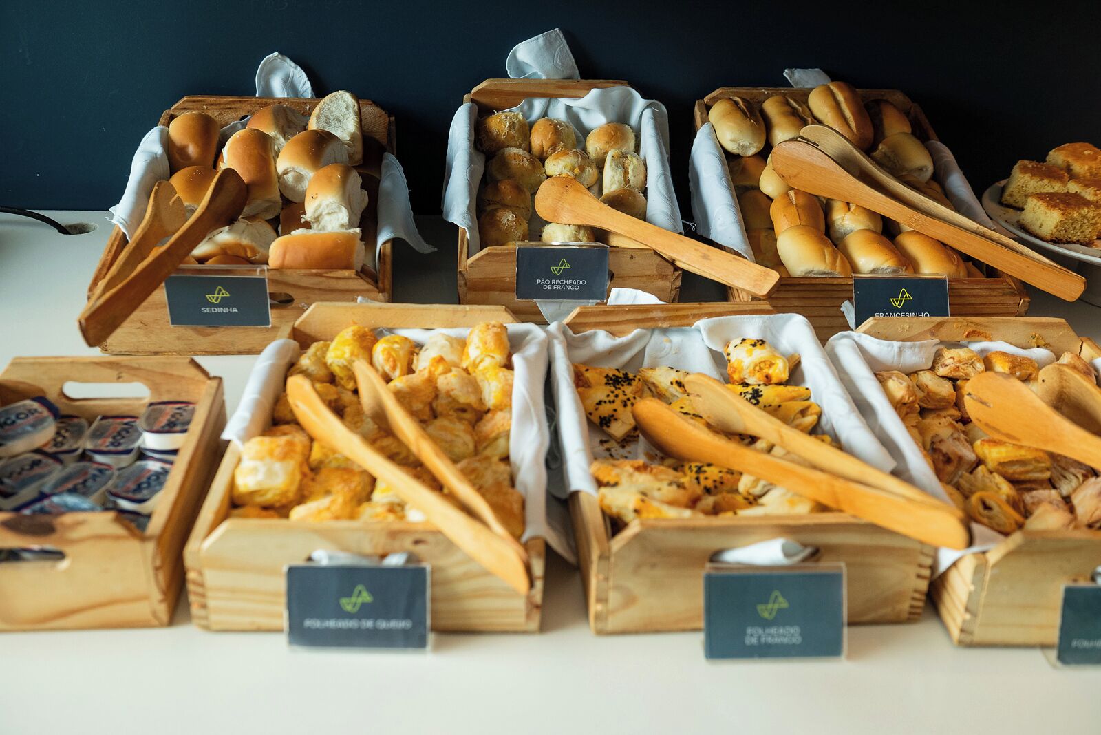 Diversos productos alimenticios expuestos en cajas de madera con etiquetas, como pan, pasteles y otras preparaciones horneadas.