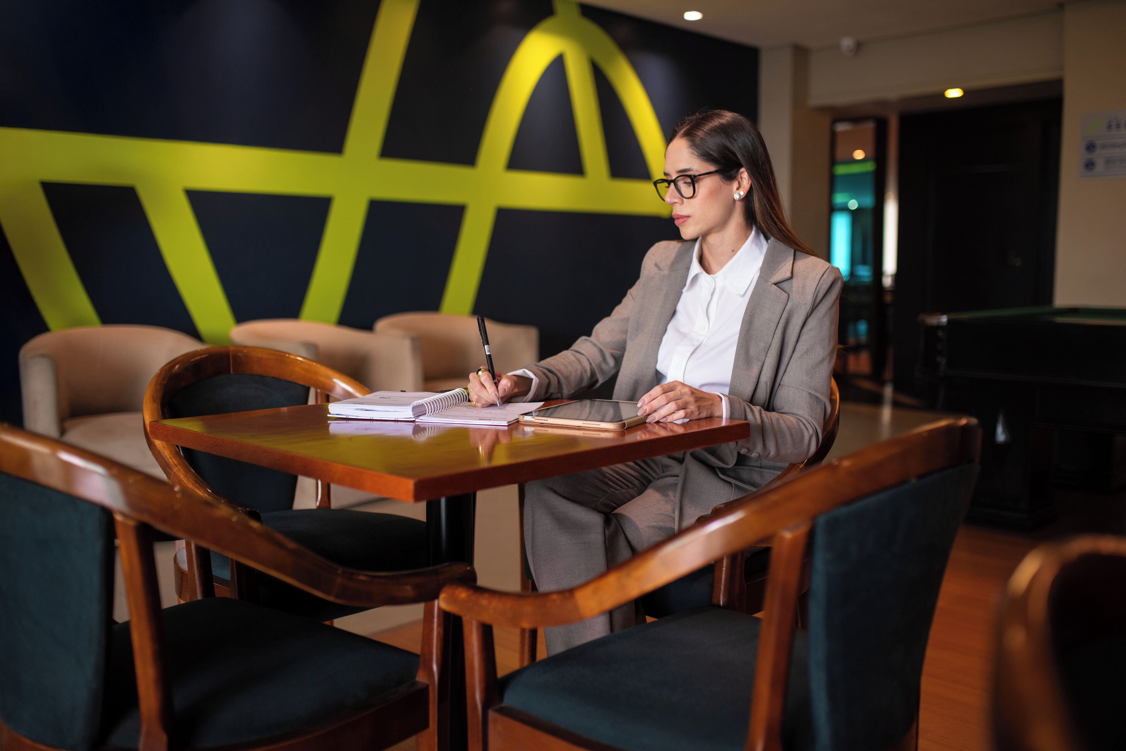Une personne est assise à une table en bois, devant un carnet et un stylo, dans une pièce dont le mur est décoré de motifs géométriques.