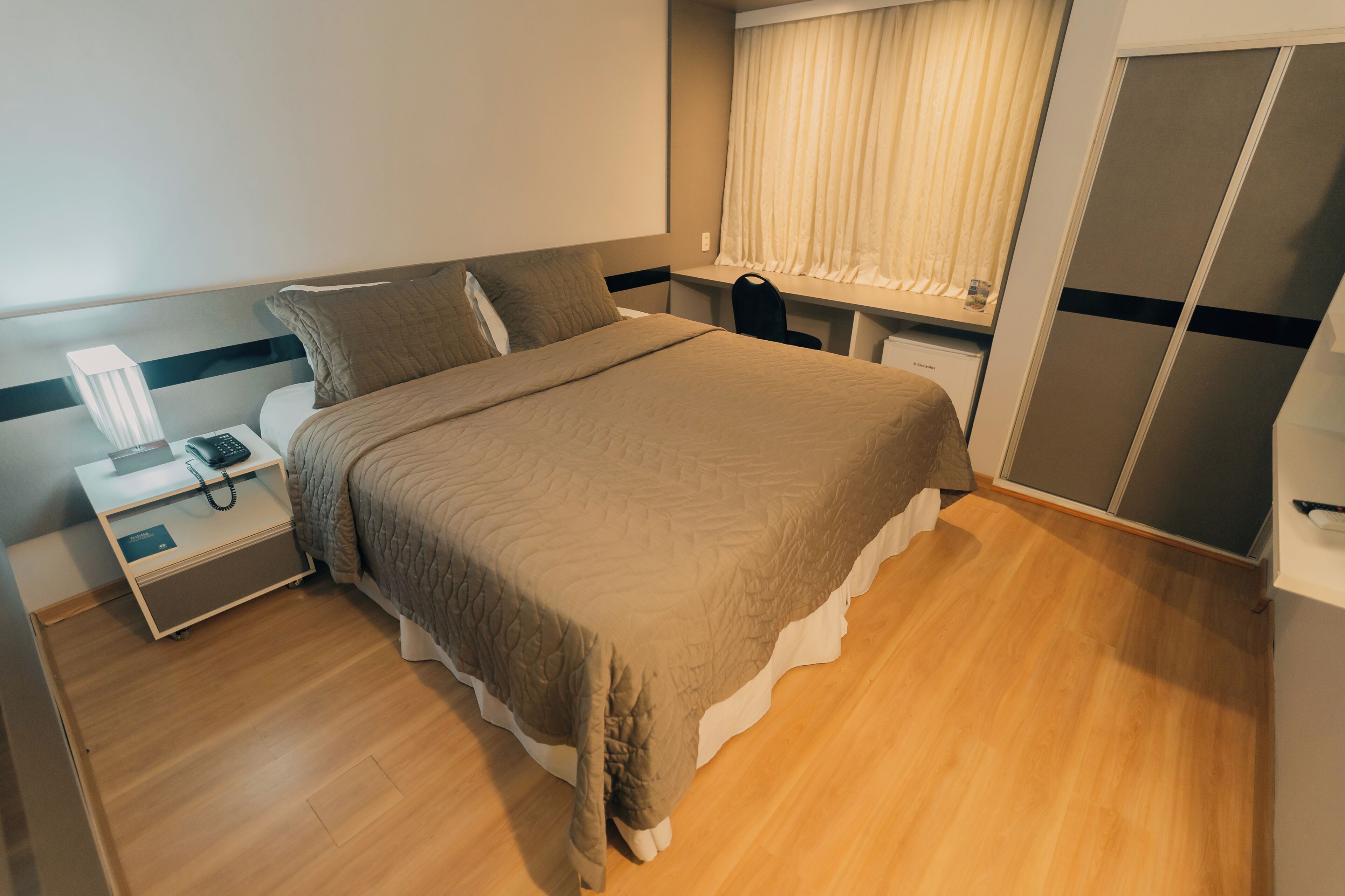 Een slaapkamer met een bed, een bureau, een stoel, een kledingkast en een raam met gordijnen.