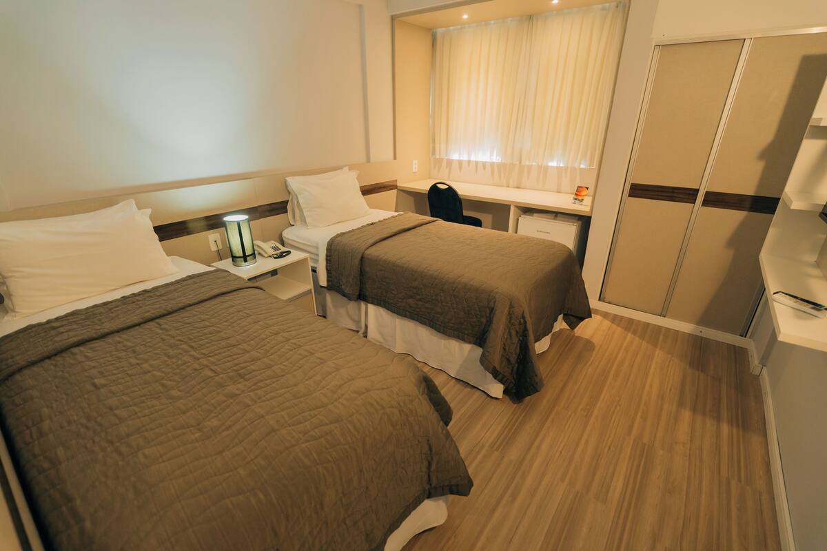 A hotel room with two beds, a desk, a chair, a wardrobe, and a window with curtains.