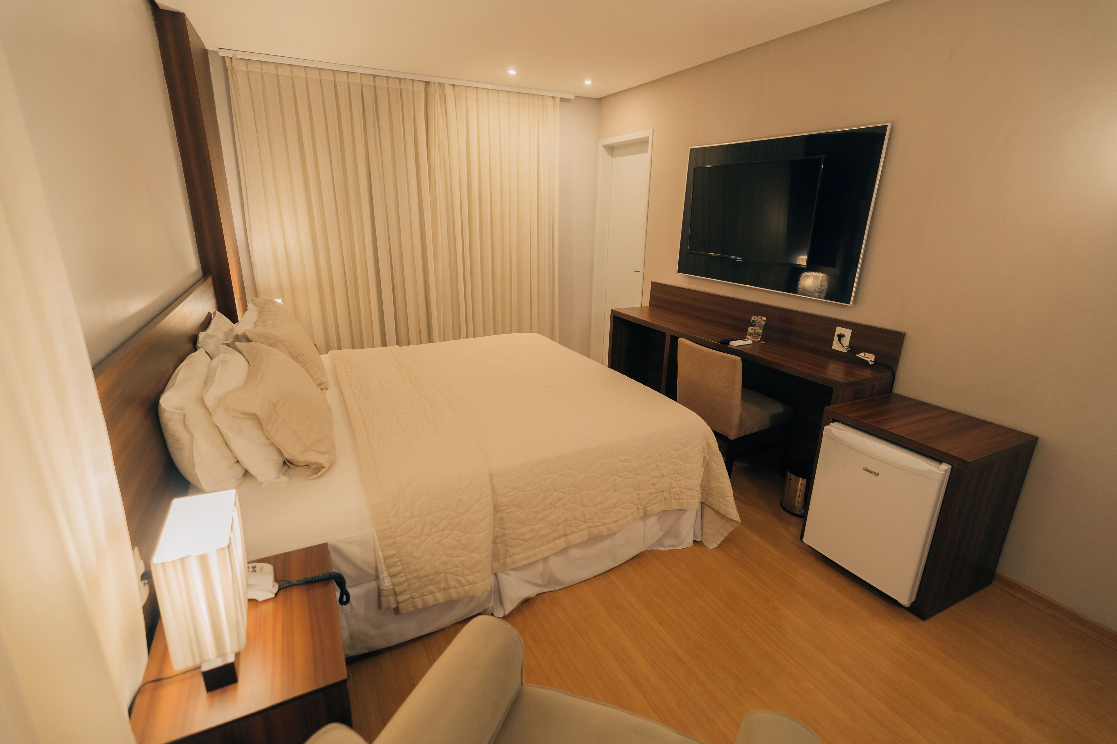 A hotel room with a bed, a desk, a small refrigerator, a TV, and a window with curtains.