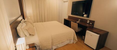 A hotel room with a bed, a desk, a small refrigerator, a TV, and a window with curtains.
