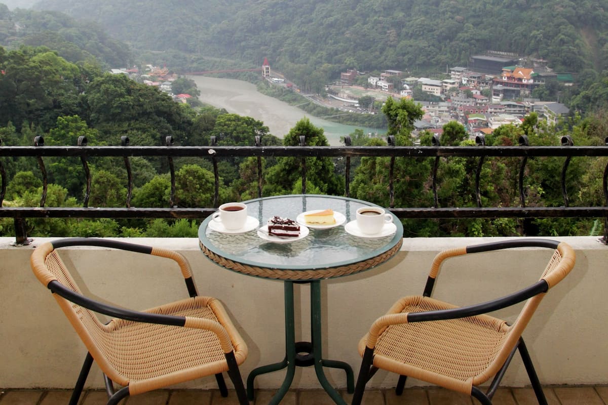 A balcony with two wicker chairs and a glass table, overlooking a river and a town with mountains in the background.