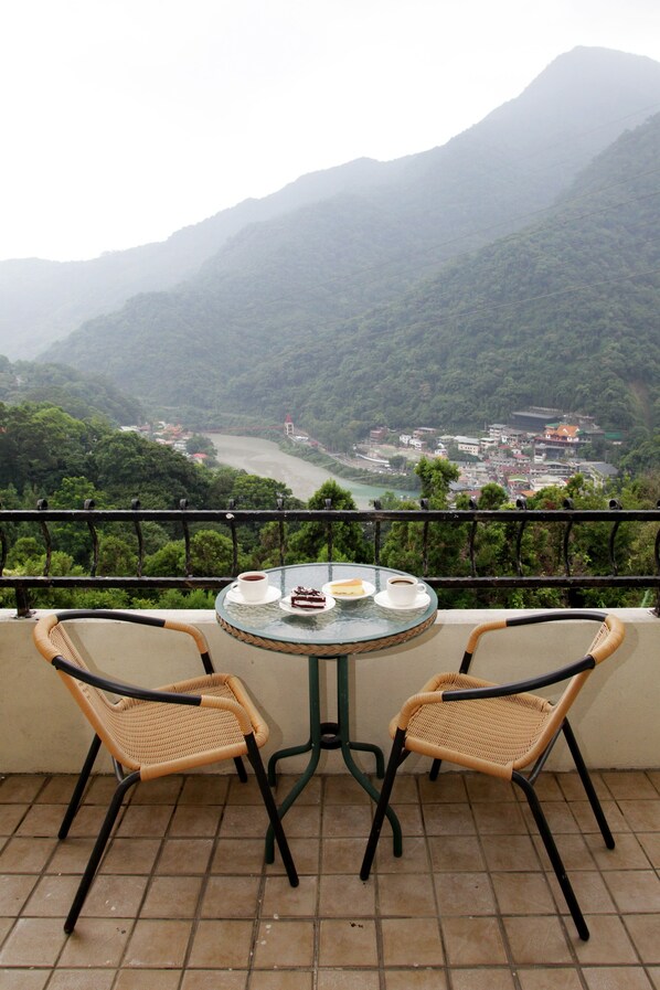 Un balcón con dos sillones de mimbre y una mesa de vidrio, con vistas a un río y un pueblo con montañas al fondo.