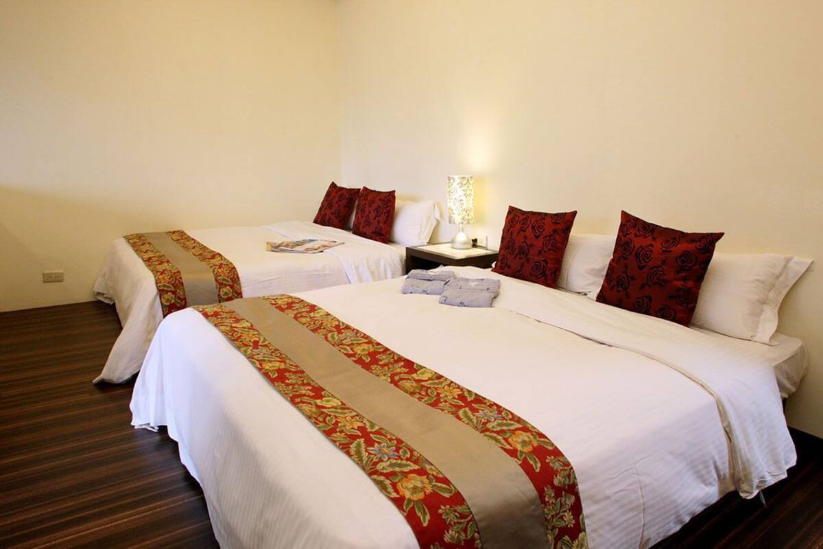 A hotel room with two beds, each with a floral bedspread and red pillows. There is a nightstand with a lamp between the beds.