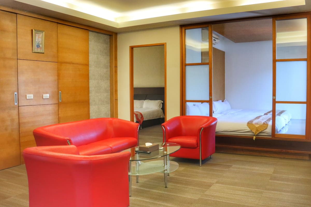 A hotel room with a bed, two red chairs, a glass coffee table, and a wooden wardrobe.
