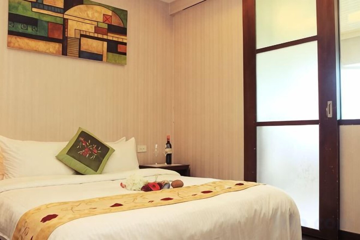 A hotel room with a bed, a nightstand, a bottle, and a window with blinds.