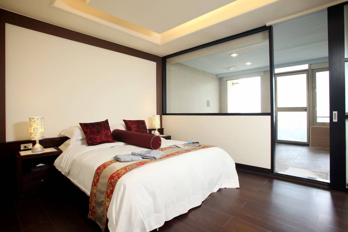 A modern hotel room with a large bed, a nightstand with a lamp, and a sliding glass door leading to a balcony.