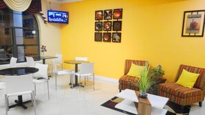 Un vestíbulo de hotel con una pared amarilla, un televisor montado en la pared, un sofá con cojines amarillos, una mesita de café y una pequeña planta.