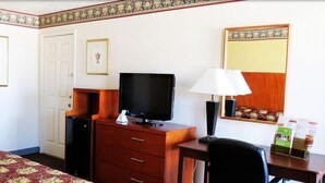 Habitación de hotel con cama, escritorio con lámpara, televisor, cómoda y un cuadro en la pared.