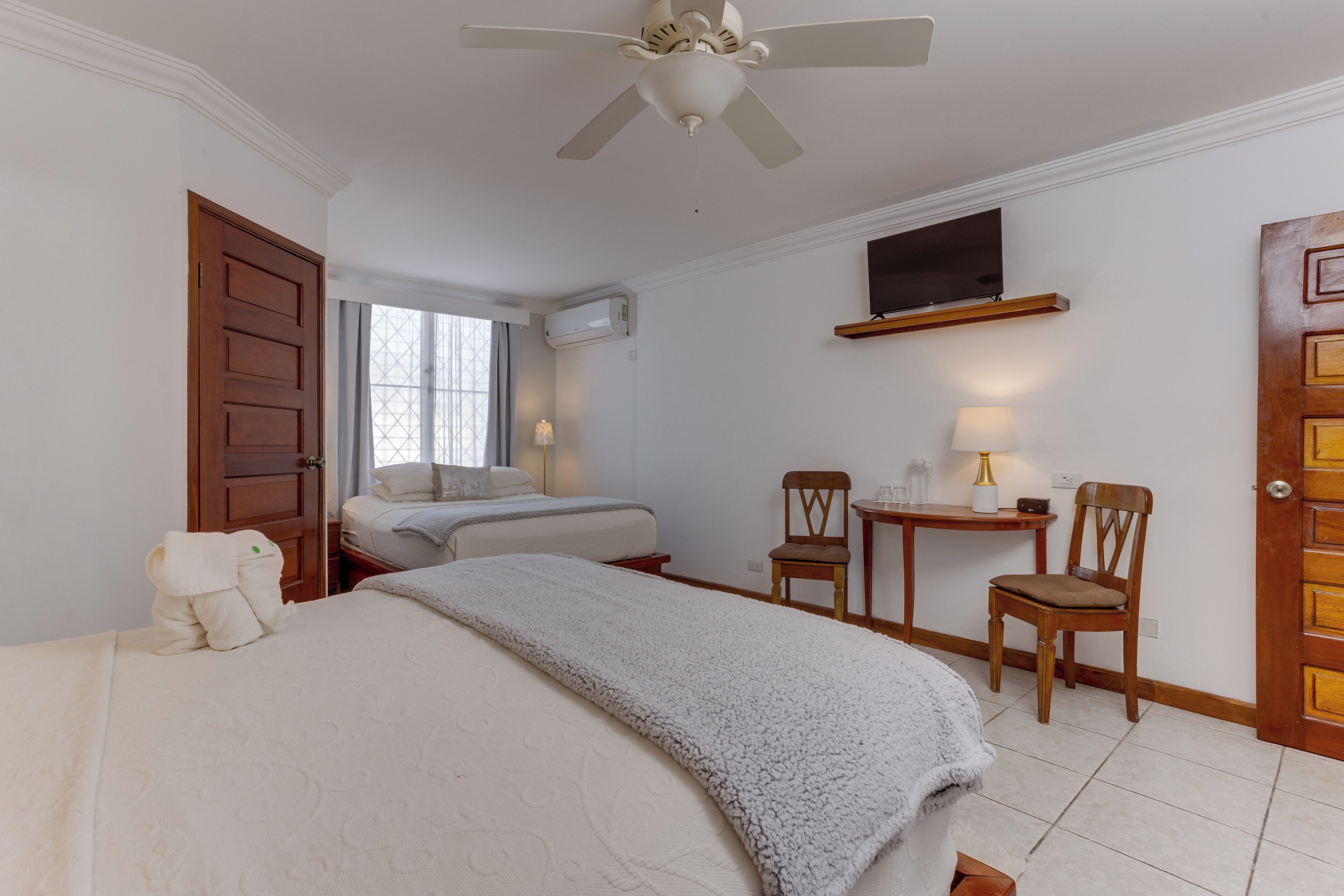A hotel room with a bed, a desk, two chairs, a television, and a ceiling fan.