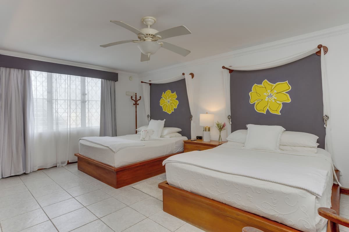 Une chambre d’hôtel avec deux lits, un ventilateur de plafond, une grande fenêtre avec des rideaux et un mur orné d’un motif floral jaune.