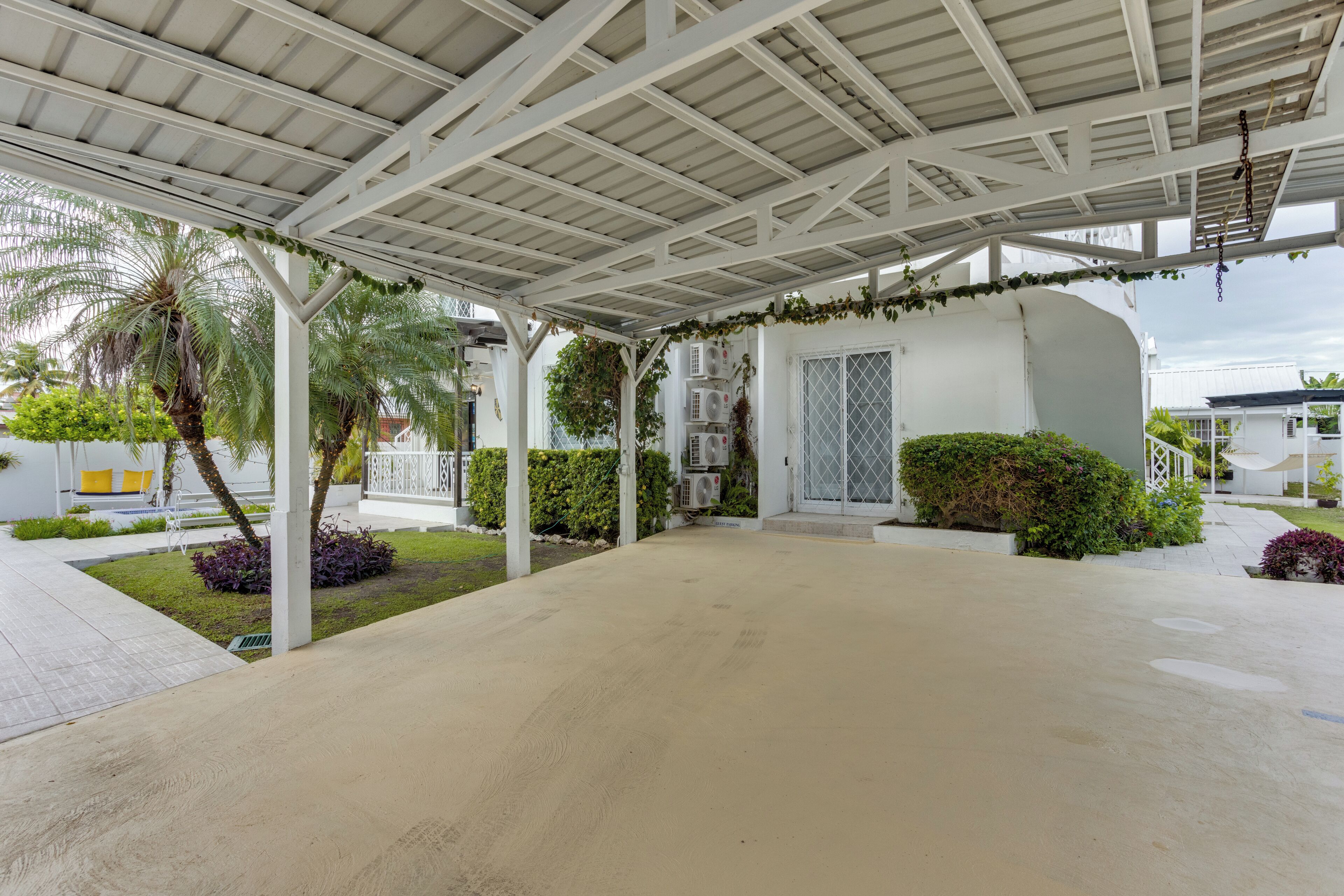 A covered outdoor area with a concrete floor, white structural beams, and greenery including palm trees and bushes.