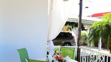 A covered outdoor patio with a table and chairs, a ceiling fan, and a view of a parked car and greenery in the background.