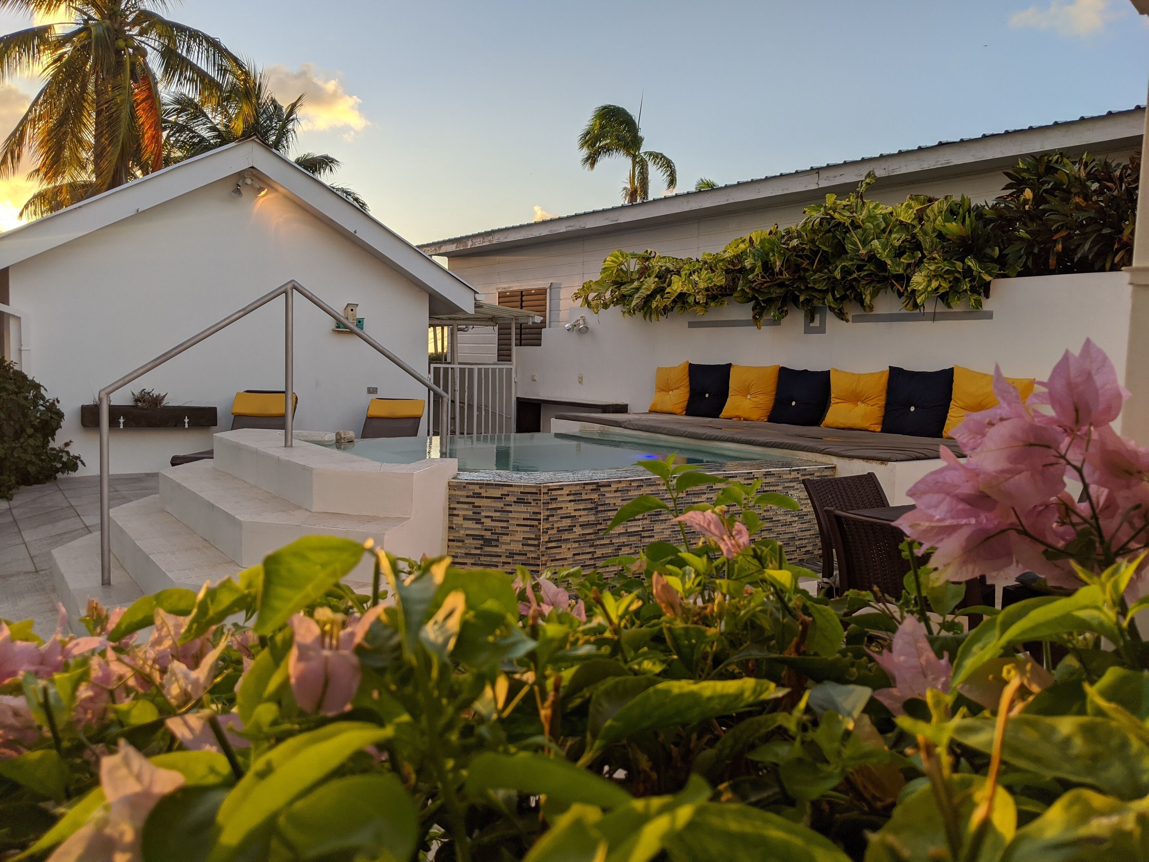 A modern outdoor pool area with a stone-tiled edge, a staircase leading to a covered seating area with yellow and black cushions, and lush greenery in the foreground.