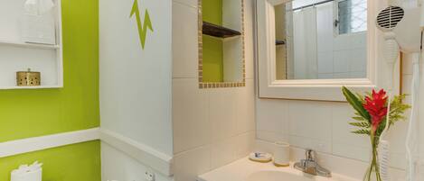 Un baño con pared verde, sanitarios blancos, un espejo, un lavamanos, un secador de pelo y un florero con flores.