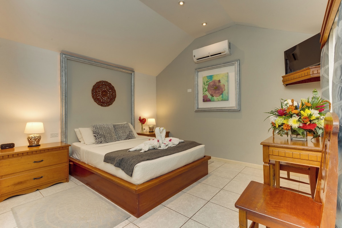 A bedroom with a wooden bed, bedside tables, a nightstand, an air conditioning unit, a framed painting, and a floral arrangement.