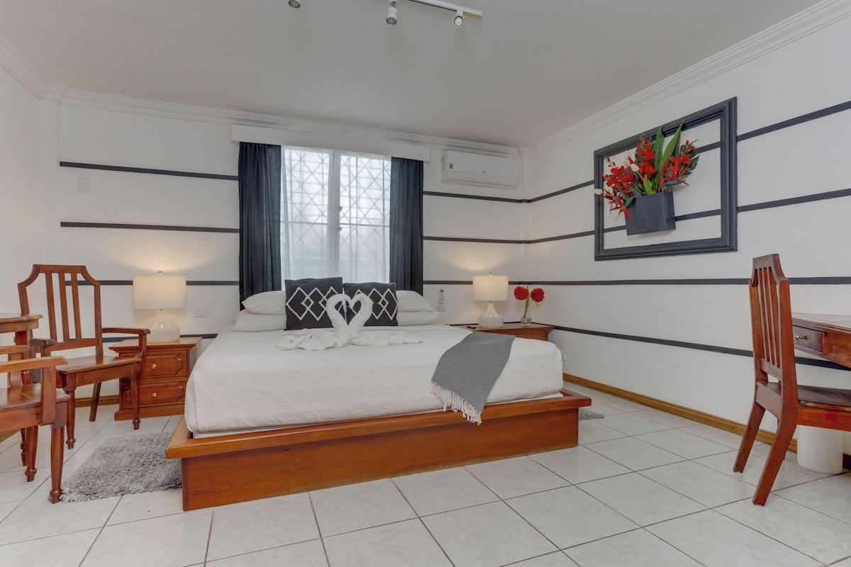 A bedroom with a bed, bedside tables, a desk, chairs, a window with curtains, and a wall-mounted air conditioner.