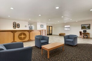 Um hall de entrada com balcão de recepção, duas poltronas azuis, uma mesa de centro de madeira e piso coberto de carpete.