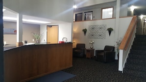 Un espace d’accueil comprenant un comptoir de réception en bois, deux fauteuils en cuir noir, une petite table avec un vase et un élément décoratif mural.