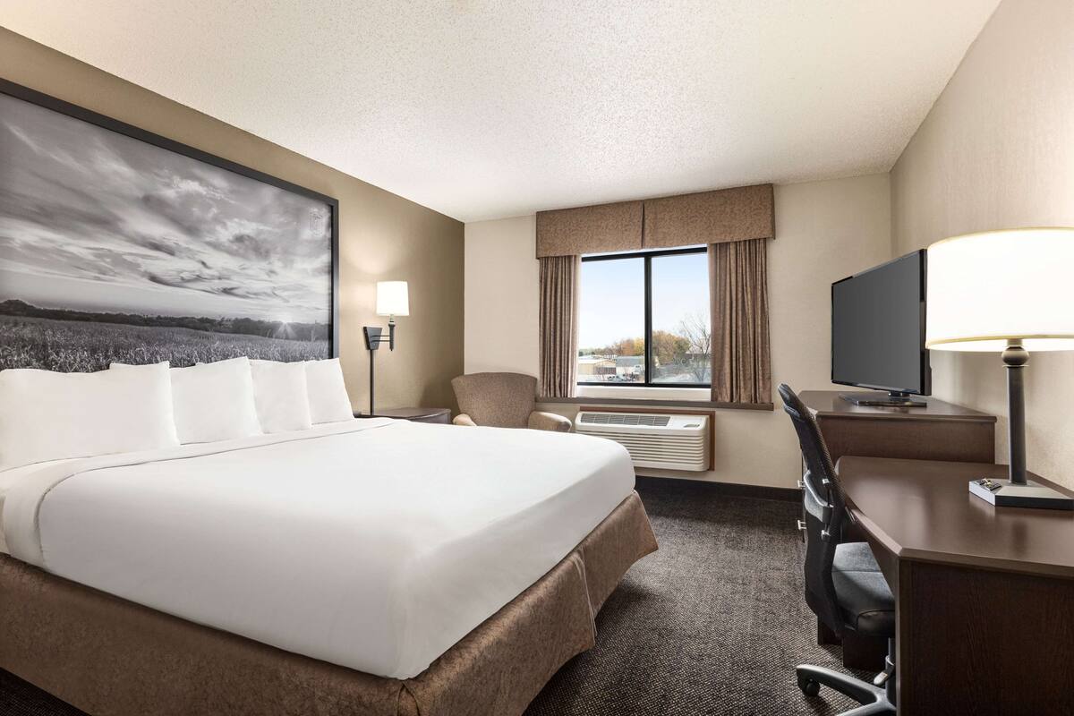 Habitación de hotel con una cama grande, un escritorio con computadora, una silla, un televisor y una ventana con cortinas.