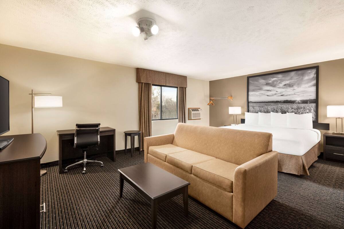 Habitación de hotel con cama, sofá, escritorio, silla, televisor y una ventana grande con cortinas.