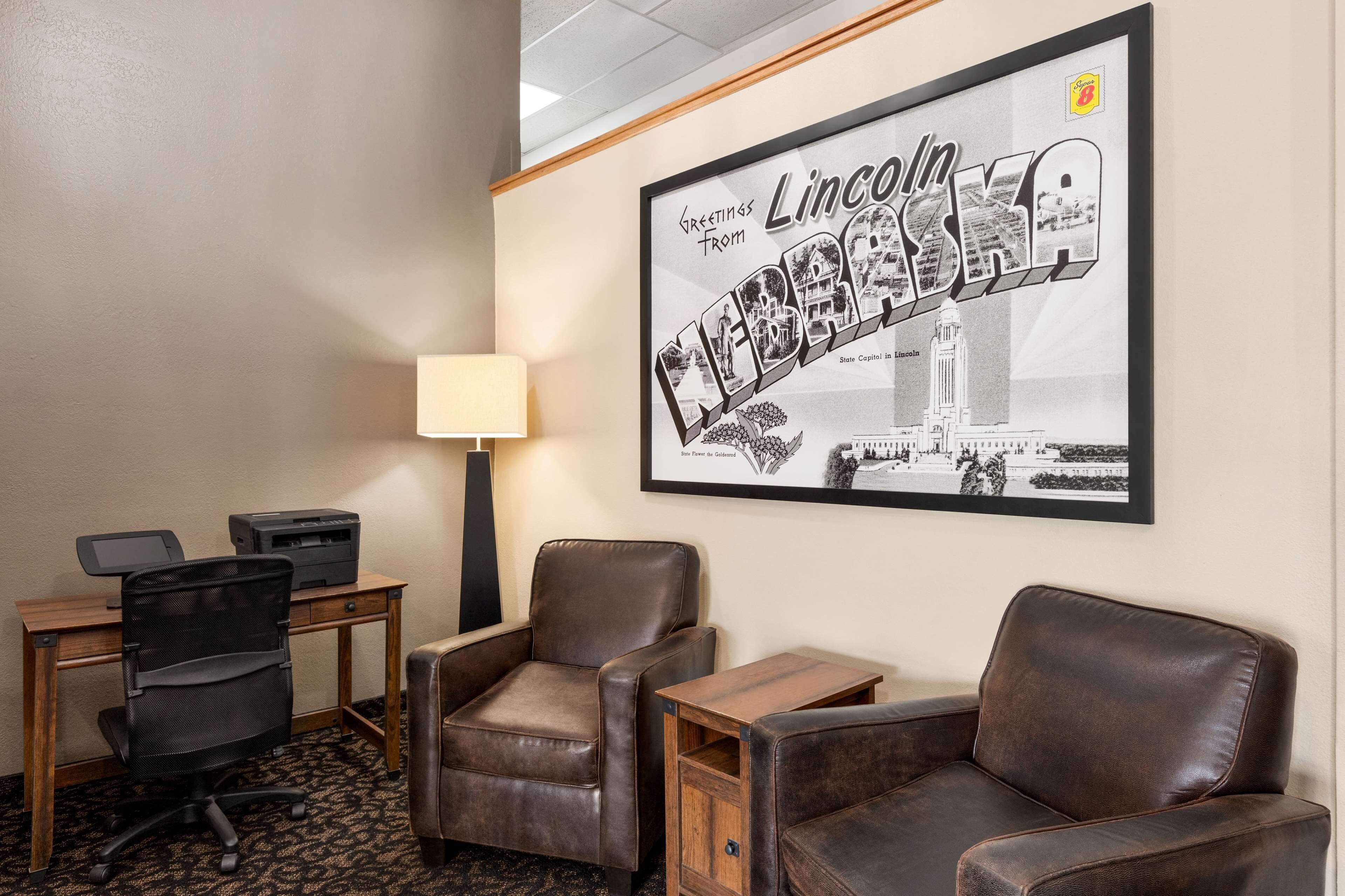 A hotel room with a desk, two leather chairs, a lamp, and a framed welcome sign.