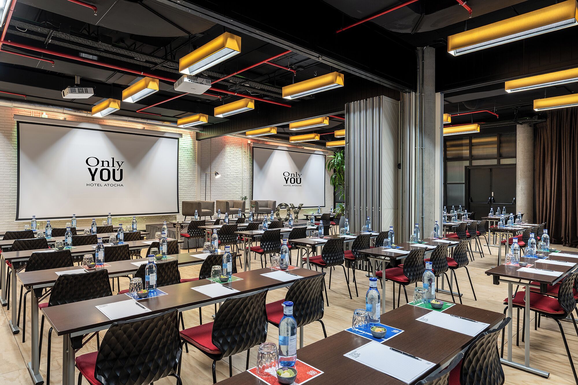 A conference room with multiple tables, chairs, and a large screen displaying 'Only YOU HOTEL ATOCHA'.