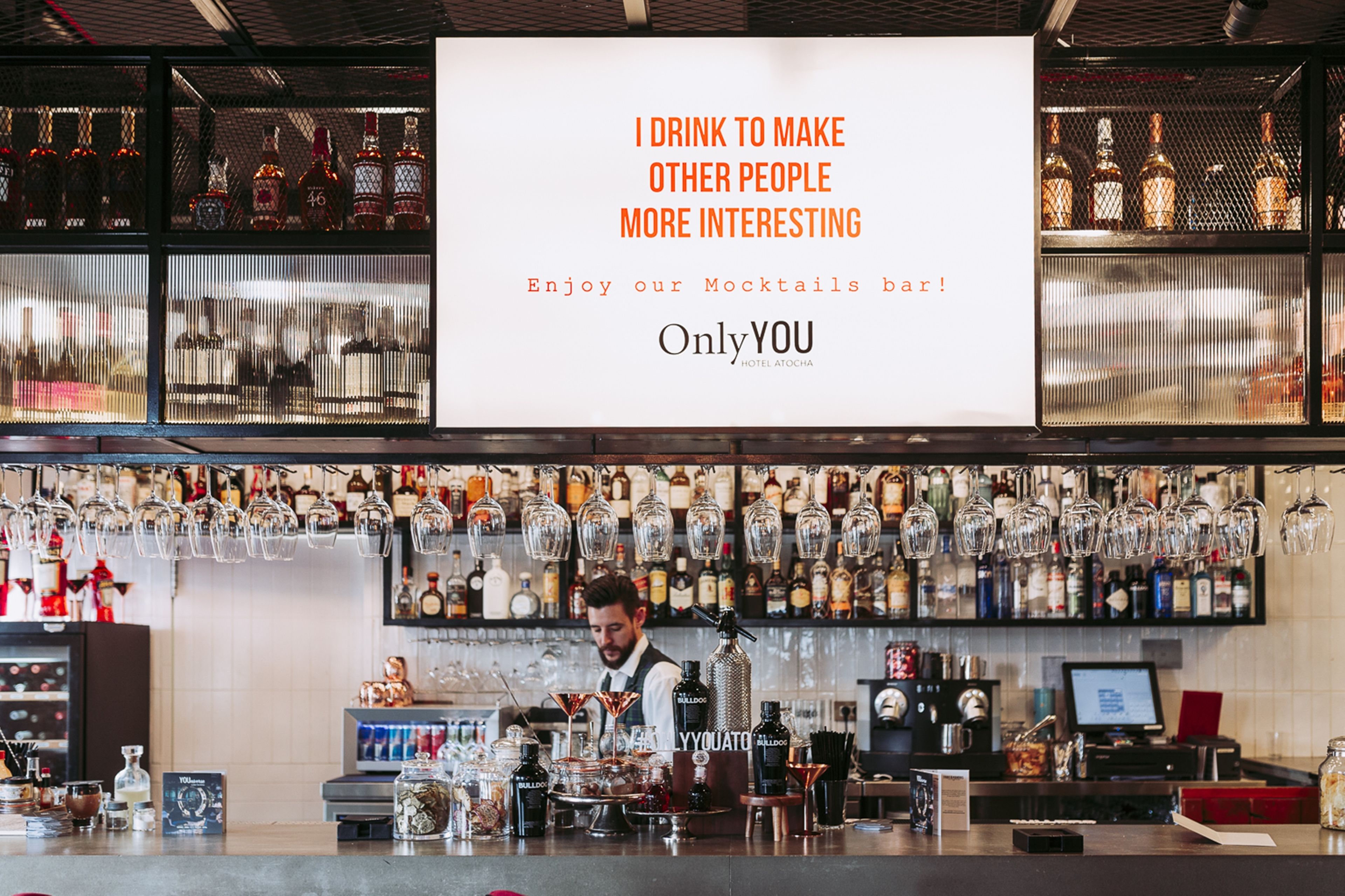 A bar area with a large sign above the counter, a bartender preparing drinks, and shelves stocked with bottles.