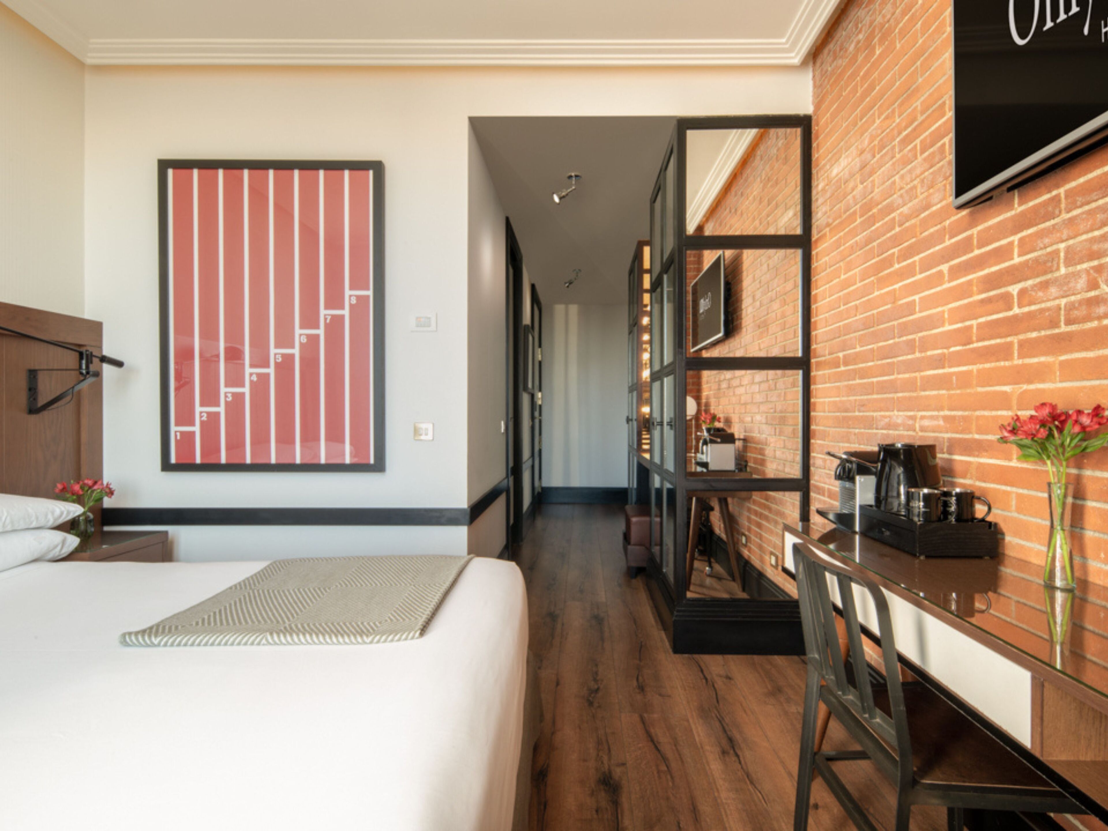 Une chambre d’hôtel avec un lit, un grand tableau abstrait, un mur en briques, un coin café et une vue sur le couloir.