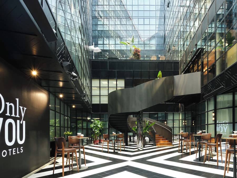 Un hall d’hôtel au design moderne, avec un sol à carreaux noir et blanc, de nombreux espaces de détente et un grand plafond en verre.