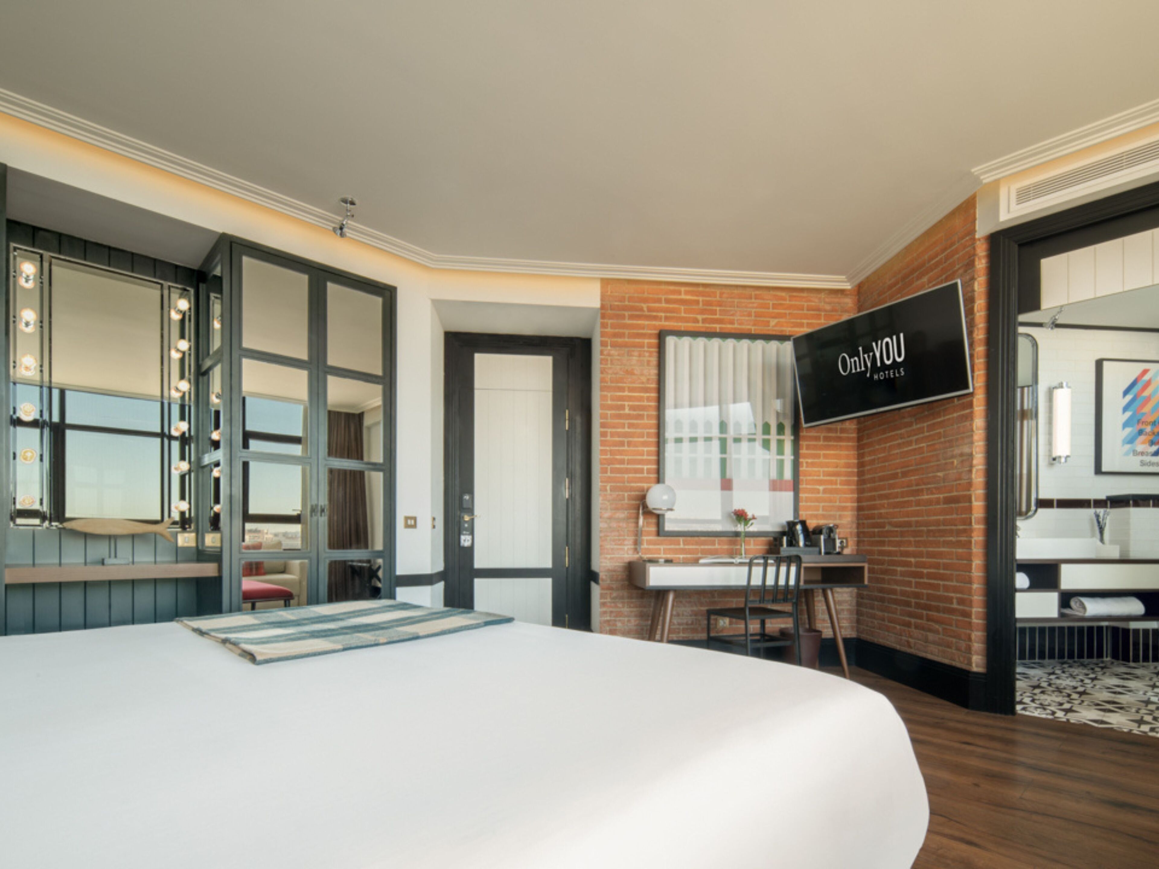 Une chambre d’hôtel comprenant un lit, un bureau, une chaise, une coiffeuse avec un miroir et une pancarte indiquant : « Only YOU Hotels ».