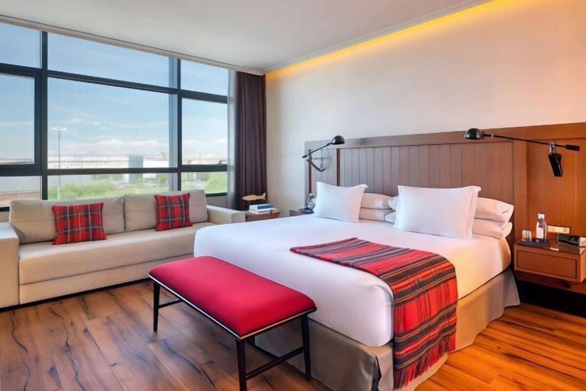 A modern hotel room with a large bed, a sofa, a bench, a nightstand, and a view of the outdoors.