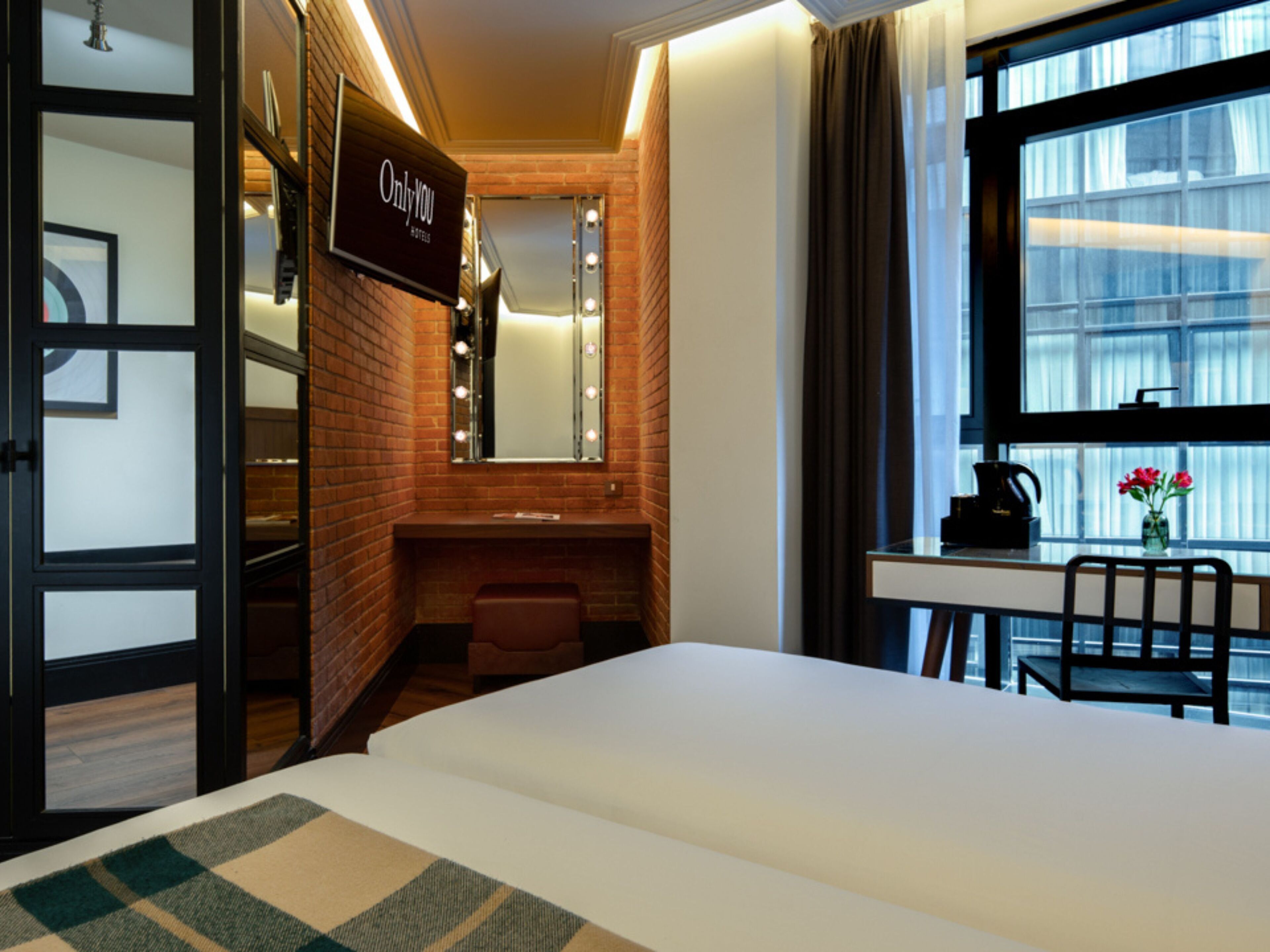 A hotel room with a bed, a desk with a chair, a vanity with a mirror and lights, and a window with curtains.