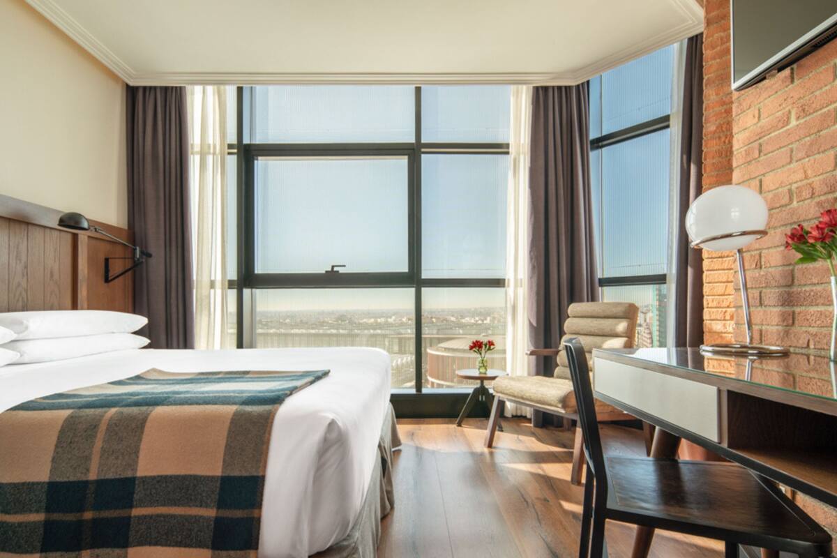 A hotel room with a large window, a bed, a desk, a chair, and a lamp.