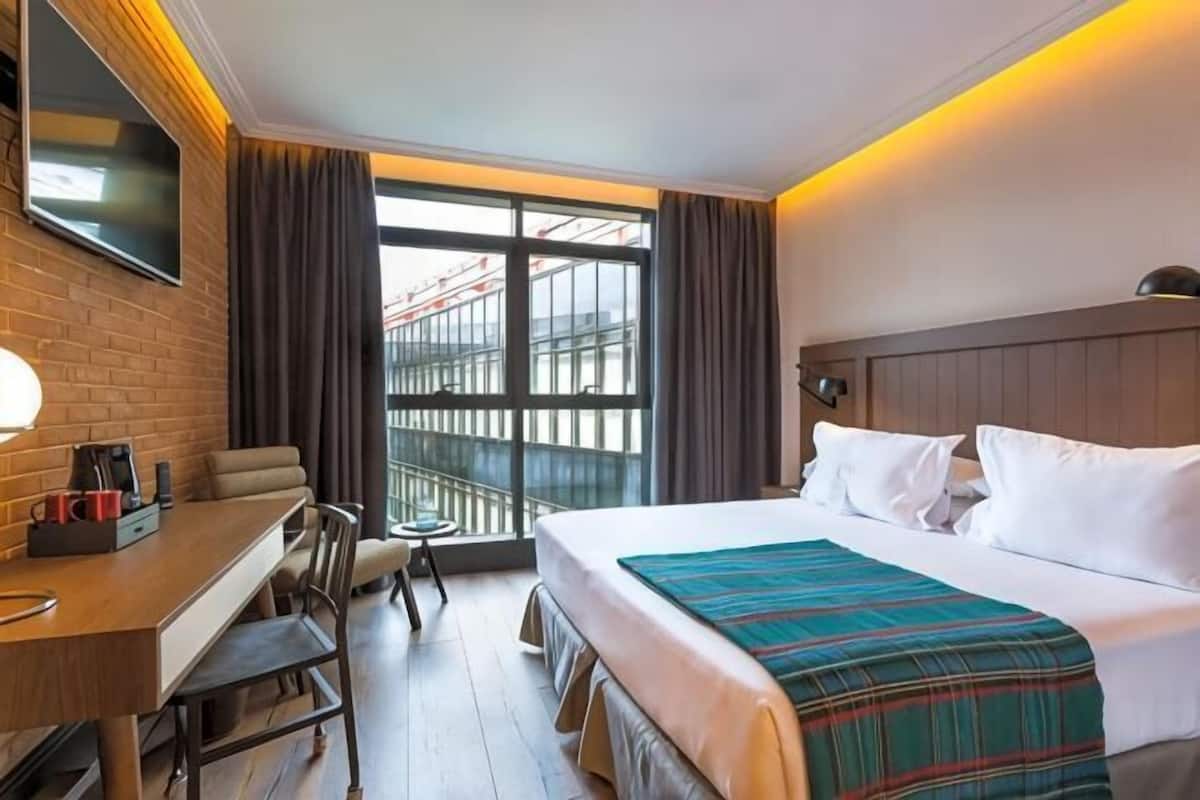 A hotel room with a bed, a desk, a chair, a TV, and a window with curtains.