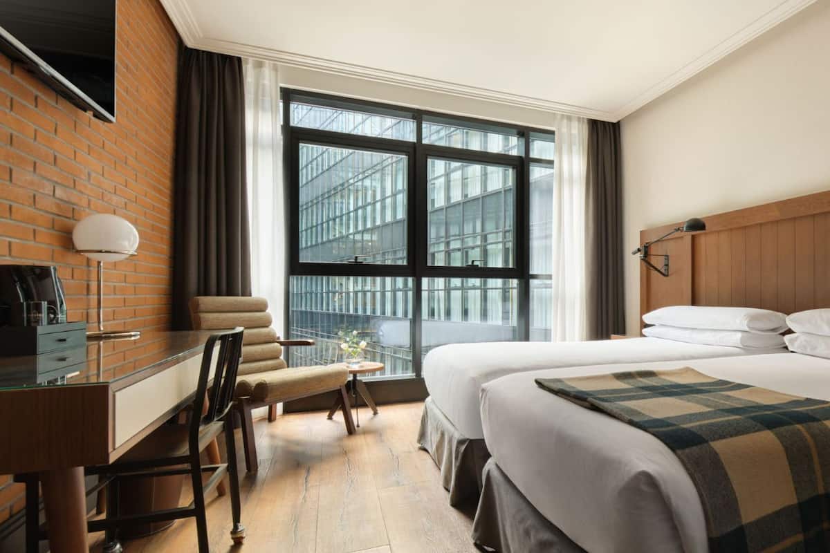 A hotel room with a desk, two beds, a large window with a view of buildings, and a brick wall.